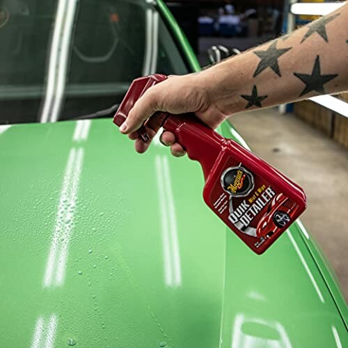 Hand holding a red quick detailer spray bottle over a green car hood.